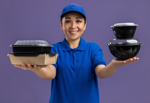 Feliz joven mujer de entrega en uniforme azul y gorra sosteniendo paquetes de alimentos sonriendo alegremente de pie sobre la pared púrpura