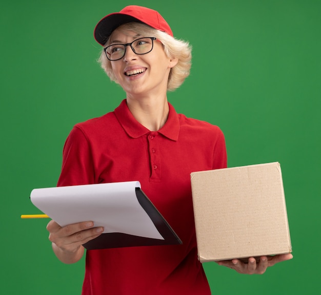 Feliz joven mujer de entrega en uniforme rojo y gorra con gafas con caja de cartón con portapapeles con páginas en blanco y lápiz mirando a un lado con una sonrisa en la cara de pie sobre la pared verde