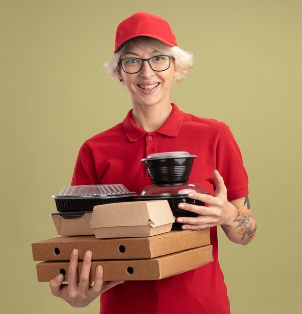 Feliz joven mujer de entrega en uniforme rojo y gorra con gafas sosteniendo cajas de pizza y paquetes de alimentos sonriendo alegremente de pie sobre la pared verde