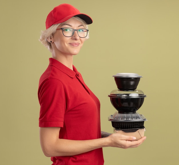 Feliz joven mujer de entrega en uniforme rojo y gorra con gafas sosteniendo la pila de paquetes de alimentos sonriendo confiados de pie sobre la pared verde