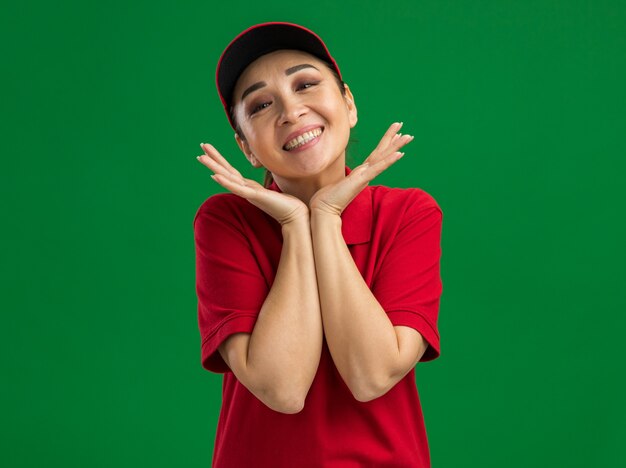 Feliz joven mujer de entrega en uniforme rojo y gorra sonriendo amigable con la mano cerca de la cara de pie sobre la pared verde