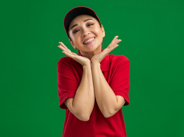 Feliz joven mujer de entrega en uniforme rojo y gorra sonriendo amigable con la mano cerca de la cara de pie sobre la pared verde