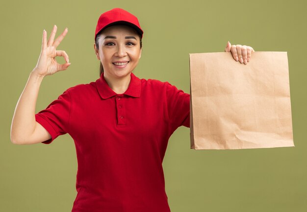 Feliz joven mujer de entrega en uniforme rojo y gorra sosteniendo el paquete de papel con una sonrisa en la cara haciendo bien firmar