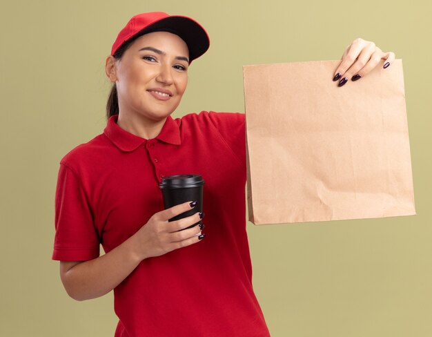 Feliz joven mujer de entrega en uniforme rojo y gorra sosteniendo el paquete de papel y la taza de café mirando al frente con una sonrisa en la cara de pie sobre la pared verde