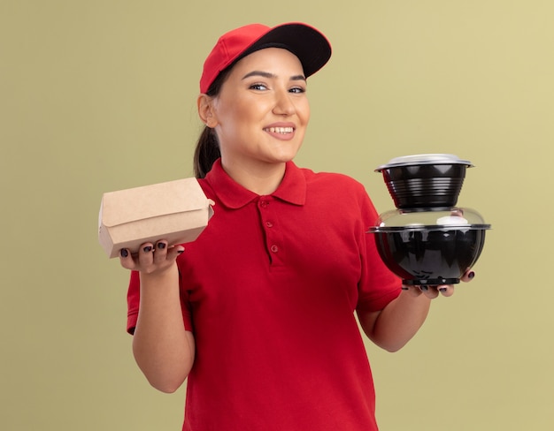 Feliz joven mujer de entrega en uniforme rojo y gorra sosteniendo paquetes de alimentos mirando al frente con una sonrisa en la cara de pie sobre la pared verde