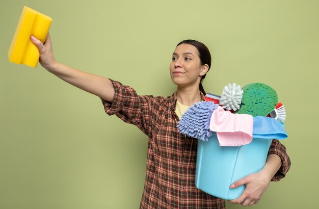 Feliz joven mujer de limpieza en camisa a cuadros sosteniendo una esponja y un balde con herramientas de limpieza mirando a un lado sonriendo confiados de pie en verde