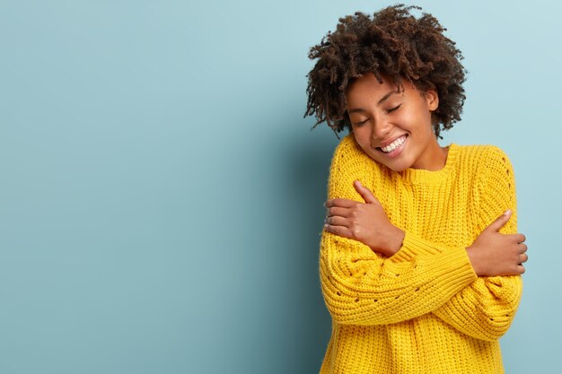 Feliz joven mujer de piel oscura se abraza a sí misma, siente un calor acogedor, viste un suéter de punto amarillo