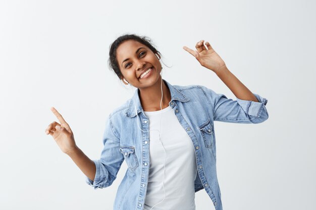 Feliz joven mujer de piel oscura con camisa azul con una sonrisa encantadora y atractiva, apuntando sus dedos hacia arriba con una mirada feliz, disfrutando de su canción favorita y bailando.