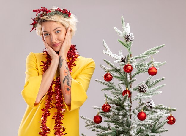 Feliz joven mujer rubia con corona de Navidad y guirnalda de oropel alrededor del cuello de pie cerca del árbol de Navidad decorado manteniendo las manos en la cara mirando a cámara aislada sobre fondo blanco
