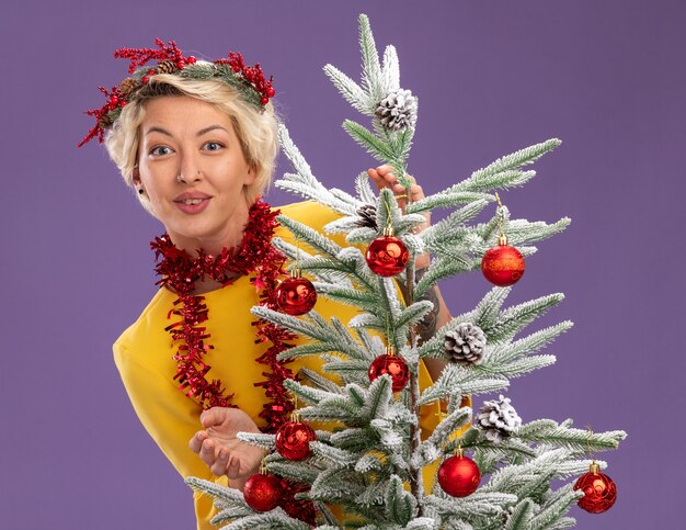 Feliz joven mujer rubia con corona de Navidad y guirnalda de oropel alrededor del cuello de pie detrás del árbol de Navidad decorado mirando aislado en la pared púrpura