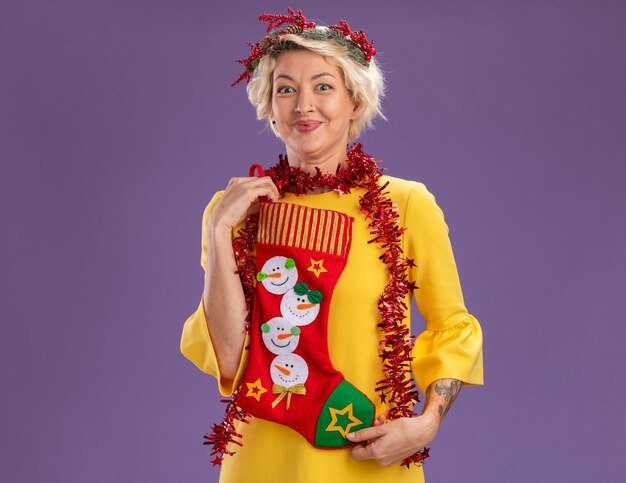 Feliz joven mujer rubia con corona de Navidad y guirnalda de oropel alrededor del cuello sosteniendo una media de Navidad mirando a cámara aislada sobre fondo púrpura con espacio de copia