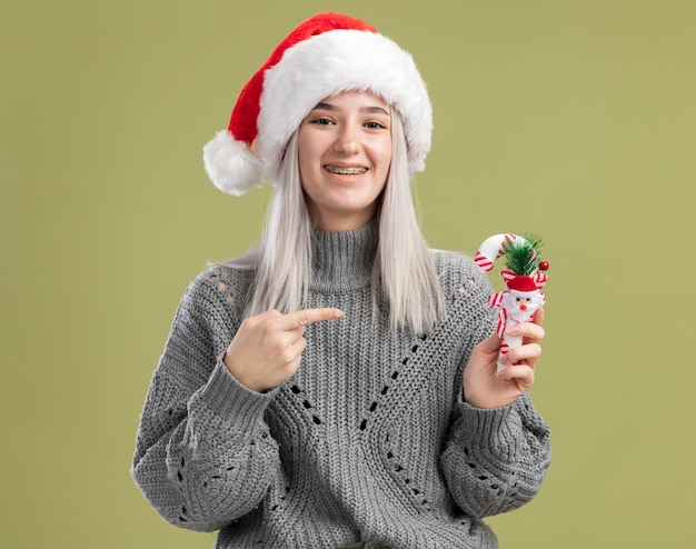 Feliz joven mujer rubia en suéter de invierno y gorro de Papá Noel con bastón de caramelo de Navidad apuntando con el dedo índice sonriendo alegremente de pie sobre la pared verde