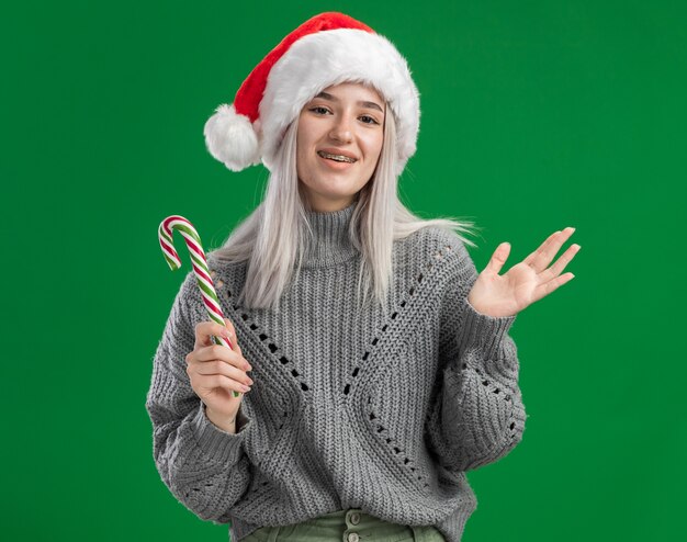 Feliz joven mujer rubia en suéter de invierno y gorro de Papá Noel con bastón de caramelo sonriendo alegremente de pie sobre la pared verde