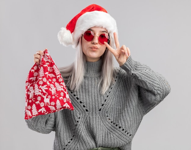 Feliz joven mujer rubia en suéter de invierno y gorro de Papá Noel con bolsa roja de santa con regalos de Navidad mostrando signo v de pie sobre la pared blanca