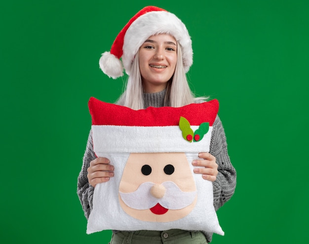 Feliz joven mujer rubia en suéter de invierno y gorro de Papá Noel sosteniendo la almohada de Navidad mirando a la cámara con una sonrisa en la cara de pie sobre fondo verde