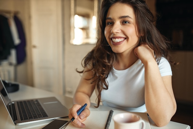 Feliz joven mujer de talla grande haciendo notas en su cuaderno mediante conexión inalámbrica a internet en la computadora portátil