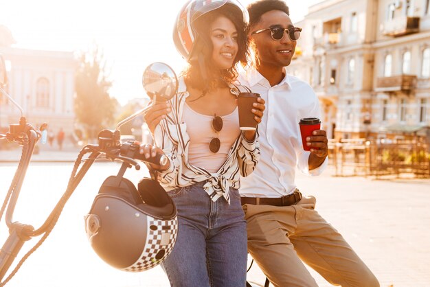 Feliz joven pareja africana tomando café mientras está de pie cerca de la moto moderna en la calle y mirando a otro lado