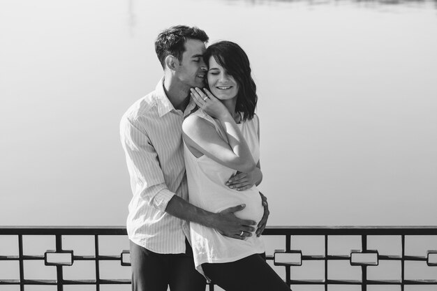 Feliz y joven pareja embarazada abrazando en la naturaleza en el lago