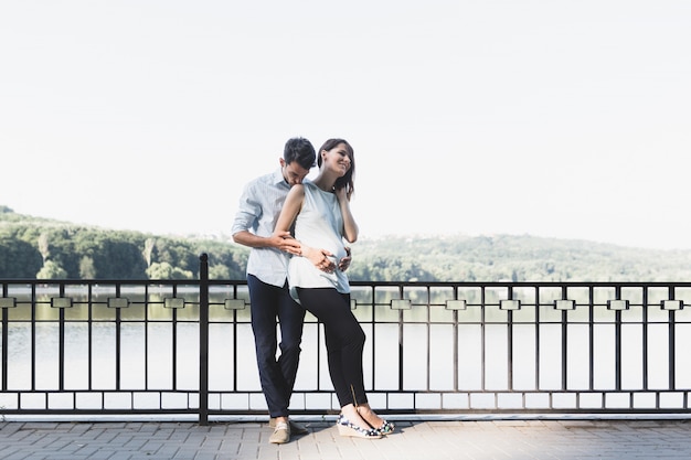 Feliz y joven pareja embarazada abrazando en la naturaleza en el lago