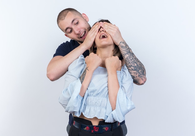 Foto gratuita feliz joven pareja hermosa de pie detrás de su novia cubriéndose los ojos sobre la pared blanca