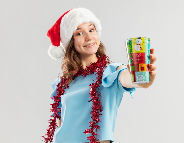 Feliz joven en la parte superior azul y gorro de Papá Noel con oropel alrededor de su cuello que muestra una colorida taza de papel sonriendo alegremente