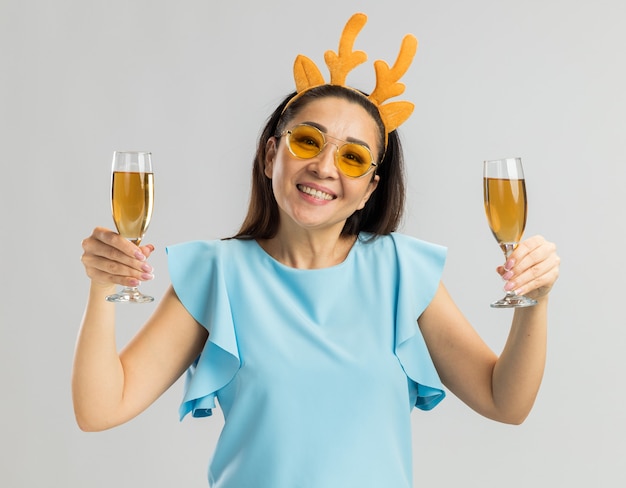 Feliz joven en la parte superior azul vistiendo un borde divertido con cuernos de ciervo y vasos amarillos sosteniendo dos copas de champán sonriendo alegremente