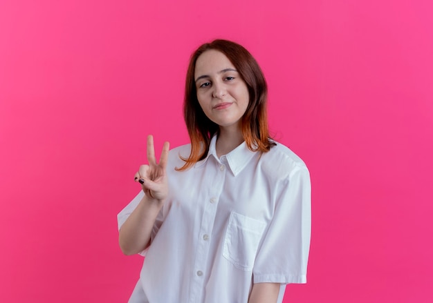 Feliz joven pelirroja mostrando gesto de paz aislado en la pared rosa