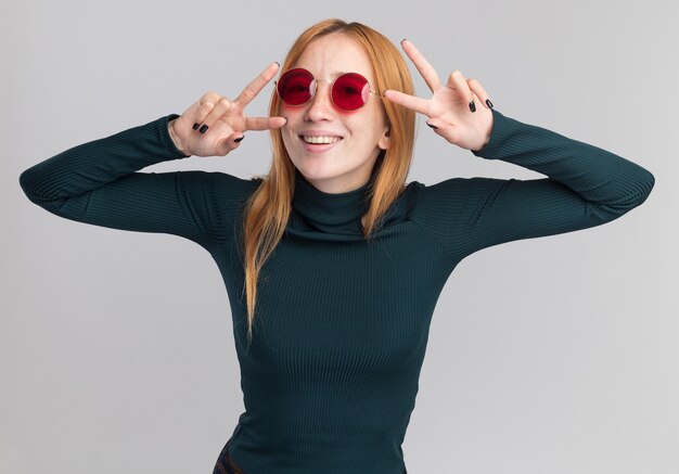 Feliz joven pelirroja pelirroja con pecas en gafas de sol gesticulando el signo de la victoria con las dos manos en blanco