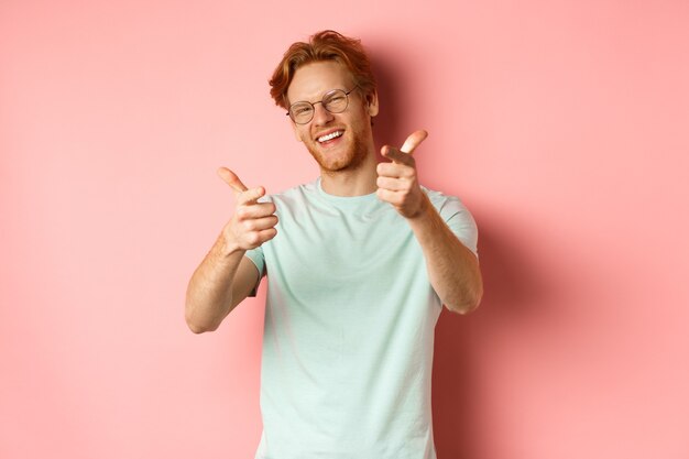 Feliz joven con pelo rojo y barba con gafas guiñando un ojo y sonriendo señalando con el dedo a vino ...