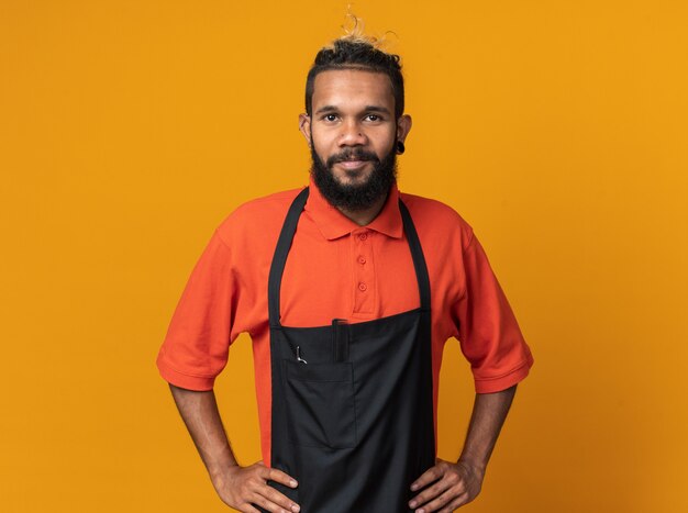 Feliz joven peluquero afroamericano vistiendo uniforme manteniendo las manos en la cintura