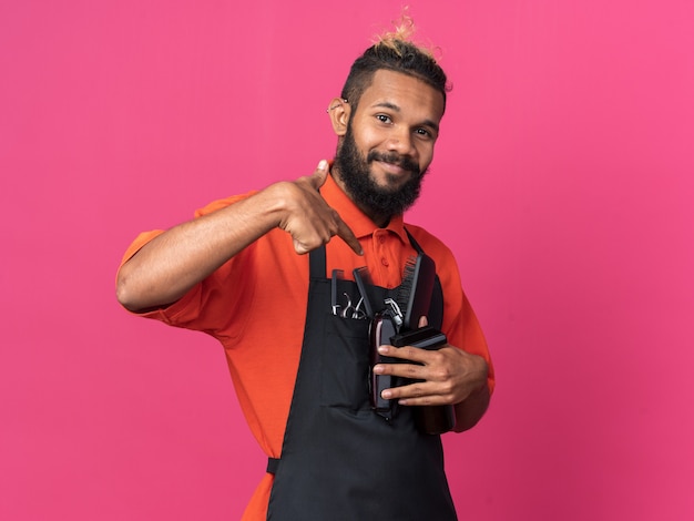 Feliz joven peluquero afroamericano vistiendo uniforme sosteniendo herramientas de peluquero apuntando a ellos