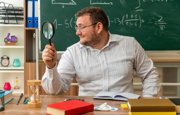 Feliz joven profesor con gafas sentado en un escritorio con útiles escolares en el aula mirando al lado a través de una lupa