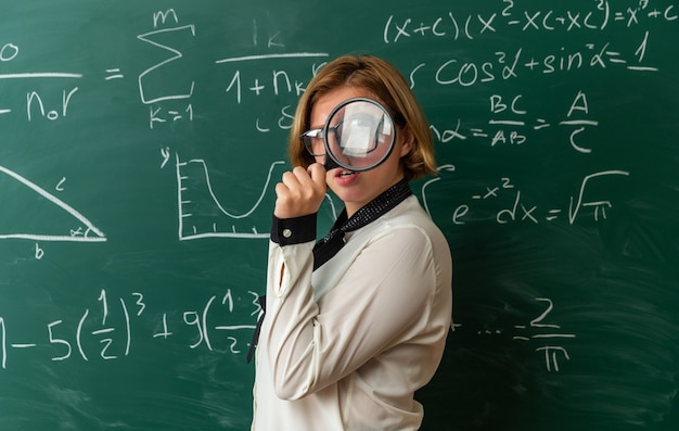 Feliz joven profesora con gafas de pie delante de la pizarra mirando a la cámara con lupa en el aula