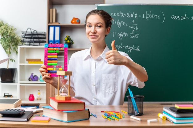 Feliz joven profesora de matemáticas sentada en un escritorio con útiles escolares sosteniendo el ábaco mirando al frente mostrando el pulgar hacia arriba en el aula