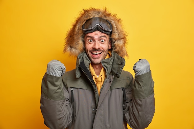 Feliz joven se regocija cuando llegó el invierno, levanta los puños cerrados, usa guantes y chaqueta con capucha