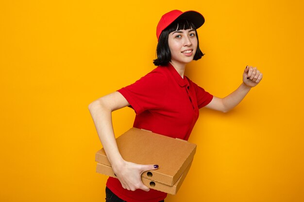 Feliz joven repartidora caucásica de pie de lado sosteniendo cajas de pizza y fingiendo correr