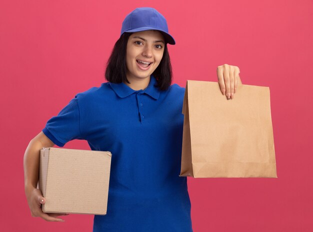 Feliz joven repartidora en uniforme azul y gorra con paquete de papel y caja de cartón sonriendo alegremente de pie sobre la pared rosa