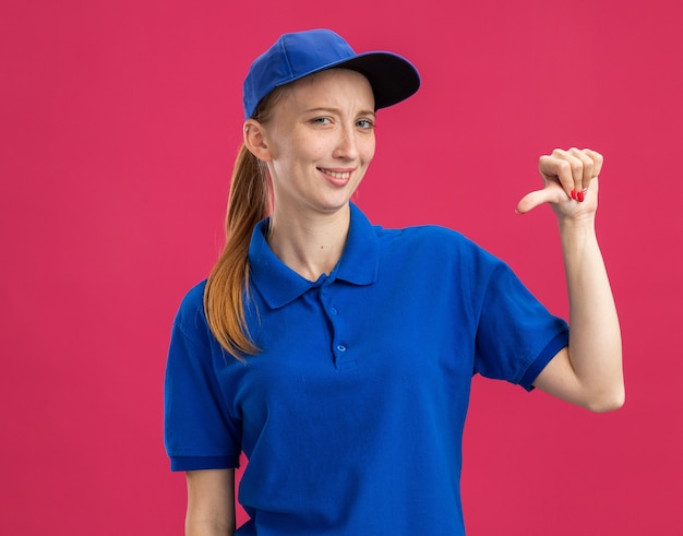 Feliz joven repartidora en uniforme azul y gorra sonriendo confiada apuntando a sí misma de pie sobre la pared rosa