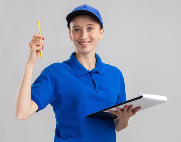 Feliz joven repartidora en uniforme azul y gorra sosteniendo lápiz y portapapeles con páginas en blanco sonriendo confiado de pie sobre la pared blanca