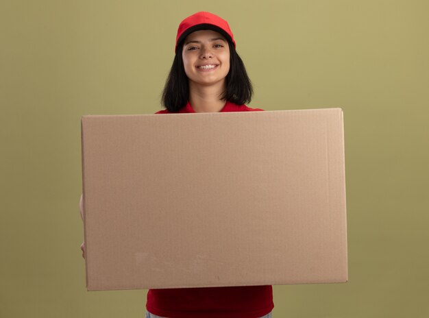 Feliz joven repartidora en uniforme rojo y gorra sosteniendo una gran caja de cartón con una sonrisa en la cara de pie sobre una pared de luz