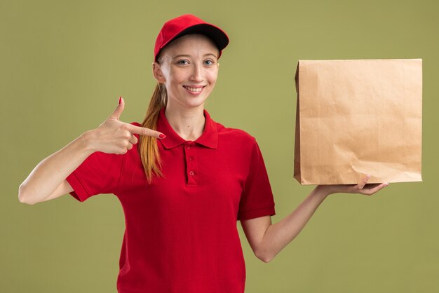 Feliz joven repartidora en uniforme rojo y gorra sosteniendo el paquete apuntando con el dedo índice sonriendo confiado