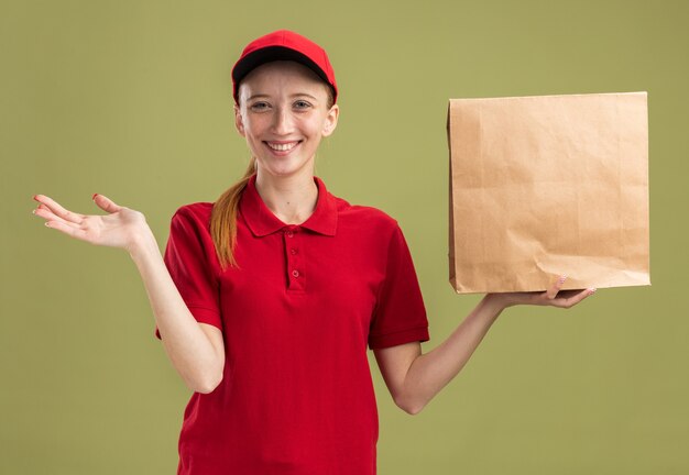 Feliz joven repartidora en uniforme rojo y gorra sosteniendo el paquete sonriendo confiado con el brazo extendido