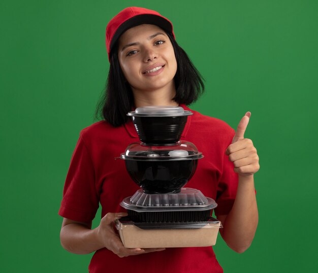 Feliz joven repartidora en uniforme rojo y gorra sosteniendo la pila de paquetes de alimentos sonriendo amigable mostrando los pulgares para arriba de pie sobre la pared verde