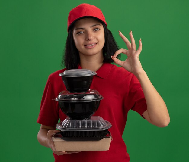 Feliz joven repartidora en uniforme rojo y gorra sosteniendo la pila de paquetes de alimentos sonriendo amistosamente mostrando signo ok de pie sobre la pared verde