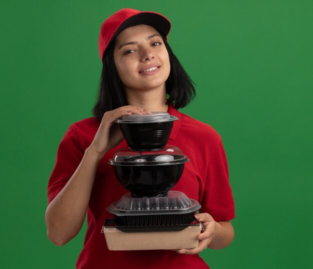 Feliz joven repartidora en uniforme rojo y gorra sosteniendo la pila de paquetes de alimentos sonriendo amistoso de pie sobre la pared verde