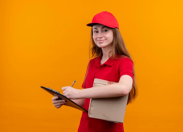 Feliz joven repartidora en uniforme rojo sosteniendo la caja y escribiendo en el portapapeles en el espacio naranja aislado