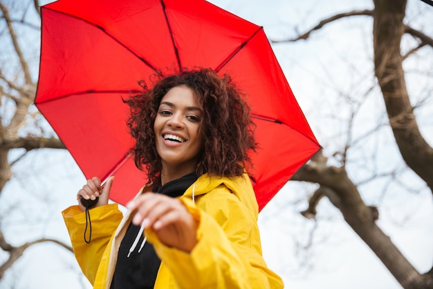 Feliz joven rizada africana con abrigo amarillo con paraguas.