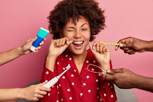 Foto gratuita feliz joven rizada se cepilla los dientes con hilo dental, se preocupa por la higiene bucal, rodeada de pasta de dientes, cepillo de dientes eléctrico y limpiador de lengua