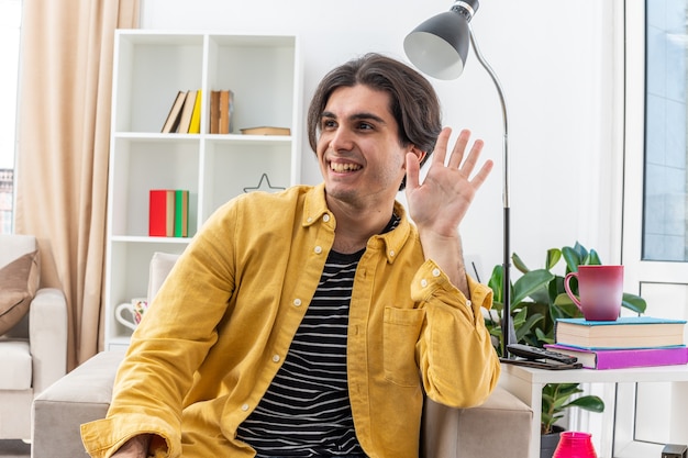 Feliz joven en ropa casual mirando a un lado saludando con la mano sonriendo alegremente sentado en la silla en la sala de luz