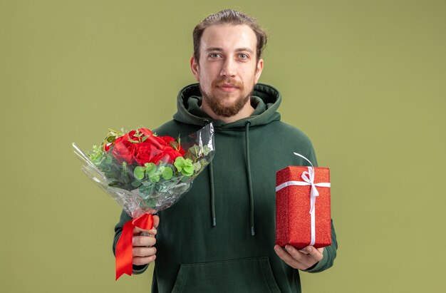 Feliz joven en ropa casual con ramo de flores y presente para su encantadora novia mirando a la cámara sonriendo confiado de pie sobre la pared verde concepto de día de San Valentín